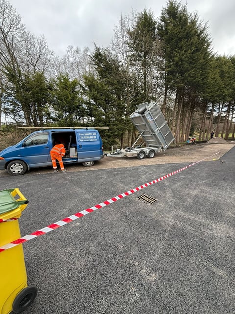 Worker in orange vest loading metal trailer from blue van on road with red and white barrier tape, surrounded by tall trees