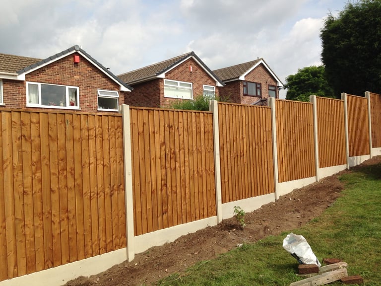 Brown wooden fence with white posts running along a residential property with brick houses and green garden visible