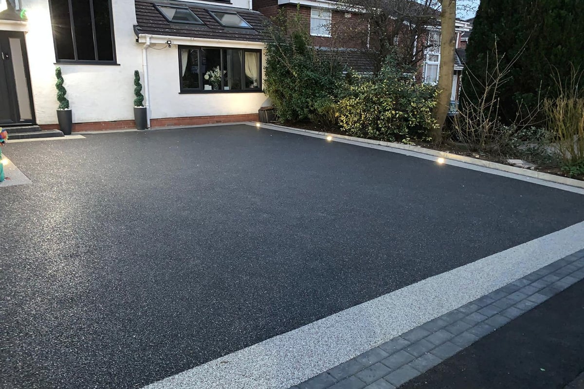 Modern residential driveway with dark asphalt center and light gray paved border, adjacent to white house with vegetation