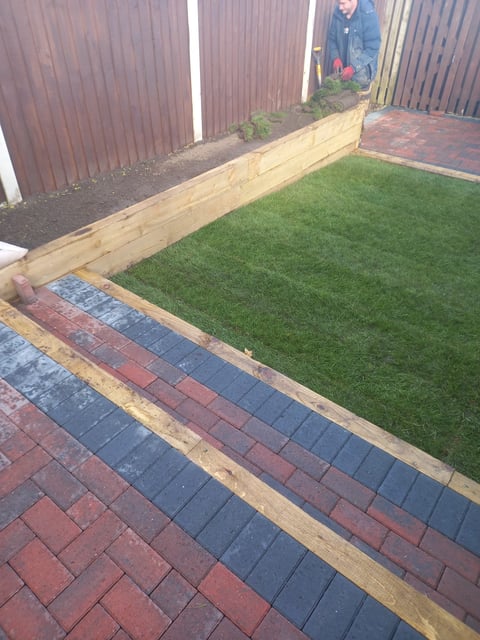 Backyard patio with red and blue brick paving transitioning to green lawn, bordered by wooden edging and corrugated metal fencing