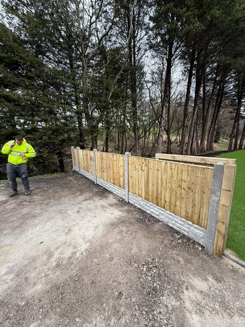 Worker in yellow safety vest stands next to newly installed wooden fence along gravel driveway with trees in background