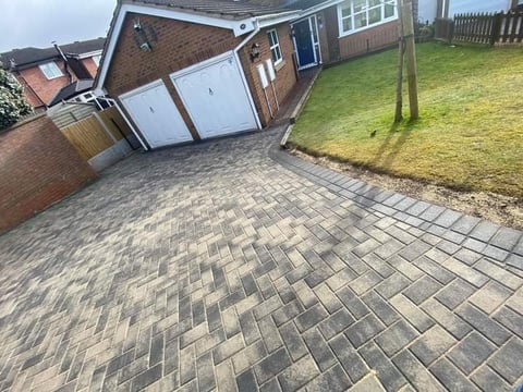 Residential driveway with patterned pavers, brick home, white garage doors, and grass lawn in background