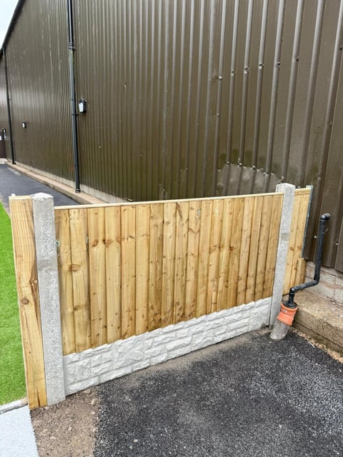 Wooden fence with concrete base posts beside a corrugated metal building wall, with green grass visible to the left