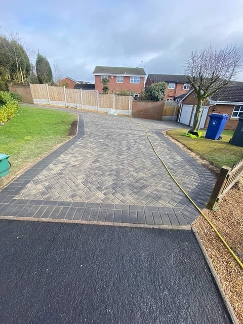 Newly installed herringbone patterned driveway with asphalt entry leading to brick-paved parking area in front of red brick house