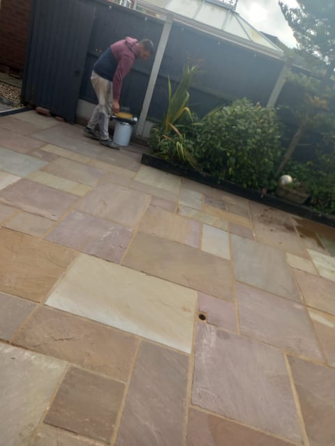 Person in maroon shirt cleaning patio with bucket near potted plants and pergola structure