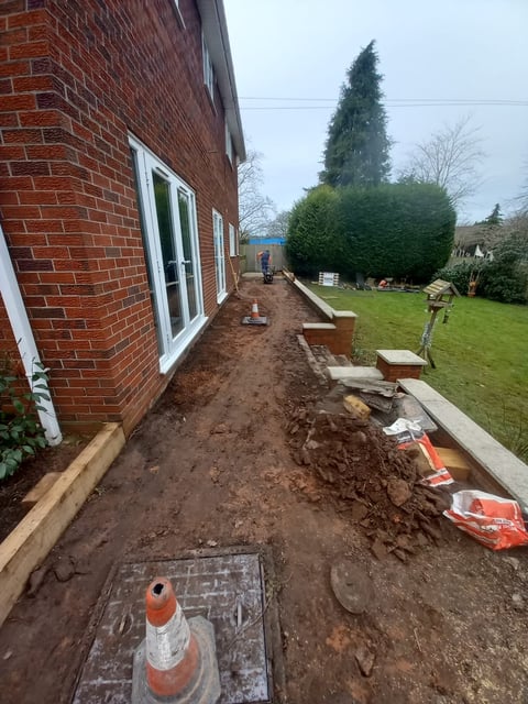 Construction area alongside brick house with exposed soil, orange traffic cones, and green lawn with landscaping in background