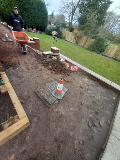 Worker in dark clothing moving soil with a red wheelbarrow during garden landscaping project with wooden borders and residential property in background