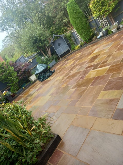 Patio garden with large stone paving, surrounded by mature trees and landscaping, featuring small garden structures
