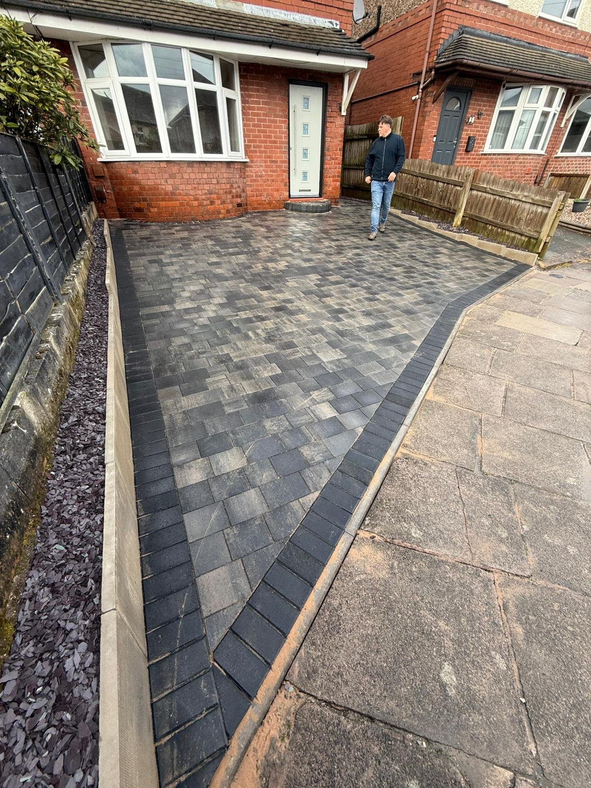 Driveway with dark patterned pavers leading to red brick house entrance with white bay window and person standing in doorway
