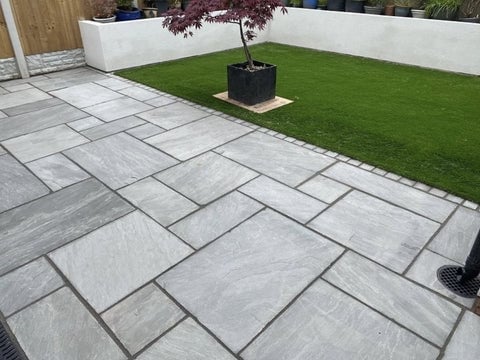 Modern patio with gray stone tiles in herringbone pattern, green lawn, black planter box with tree, and white walls