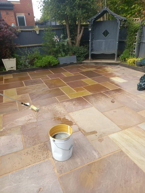 Backyard patio with freshly laid sandstone pavers in various shades, a bucket of sealant, ivy-covered walls, and a black garden shed