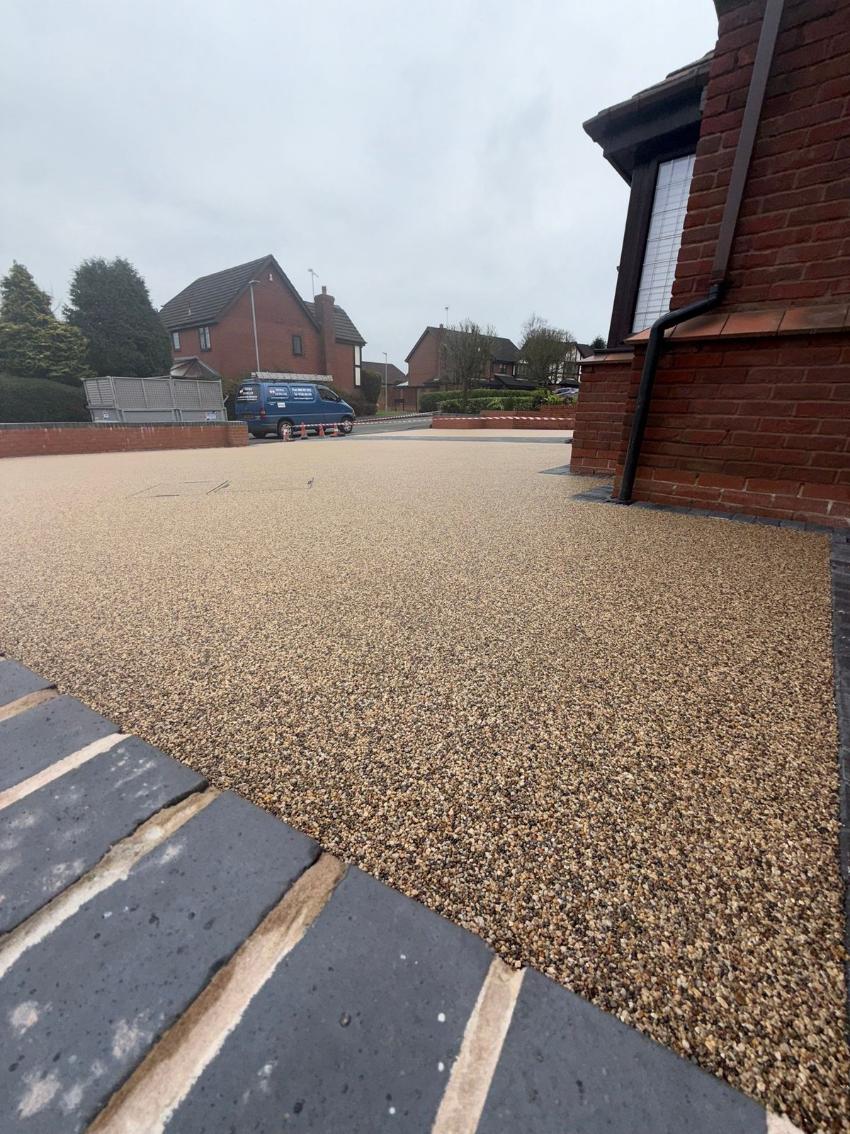 Residential driveway with gravel surface, red brick houses, and parked car in a suburban neighborhood on an overcast day