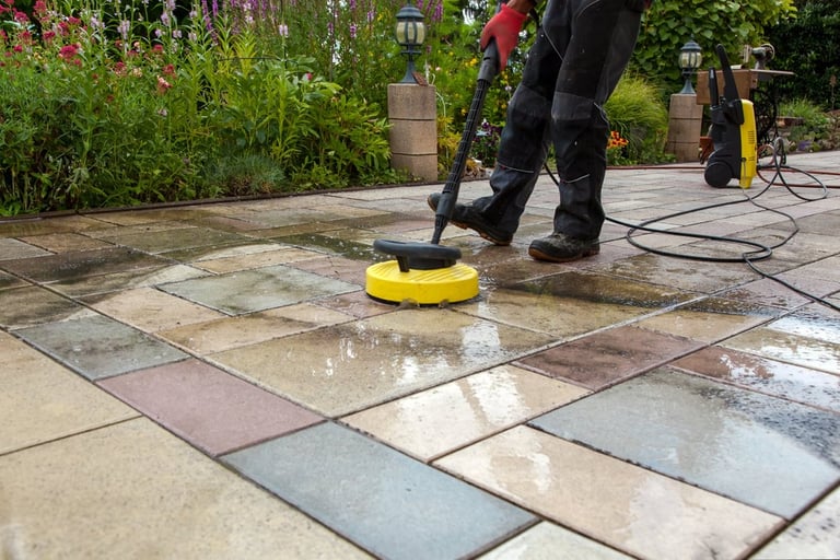Person using pressure washer with yellow attachment to clean wet patio tiles in a garden setting