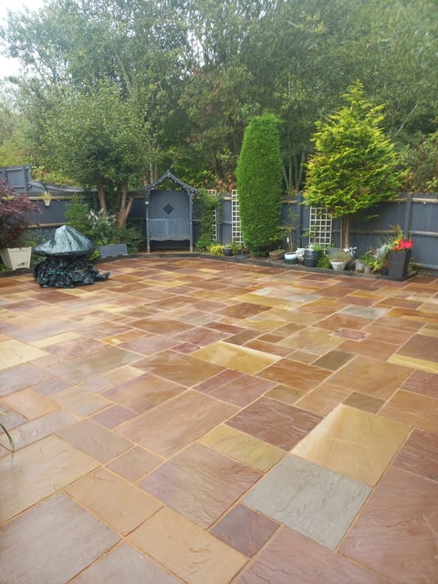 Spacious patio with terracotta and stone tiles, surrounded by lush green trees and garden structures in background