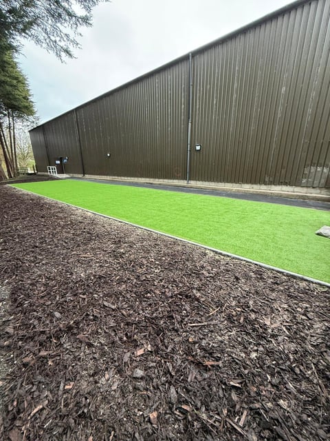 Large corrugated metal warehouse building with bright green artificial turf and dark mulch ground alongside trees