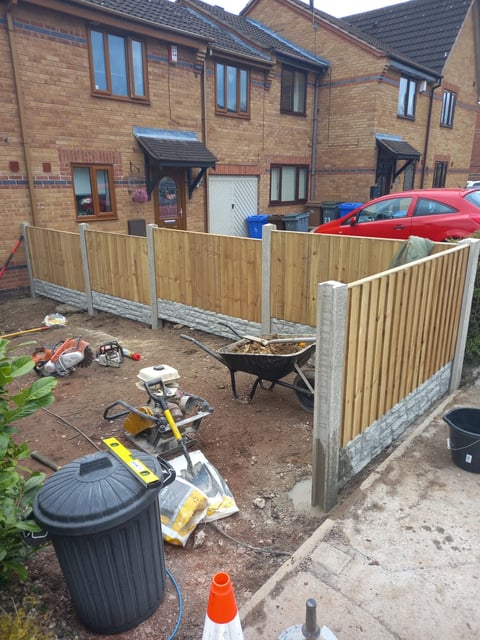 Construction site in residential yard with wooden fence panels, tools, wheelbarrow, and rubble bin
