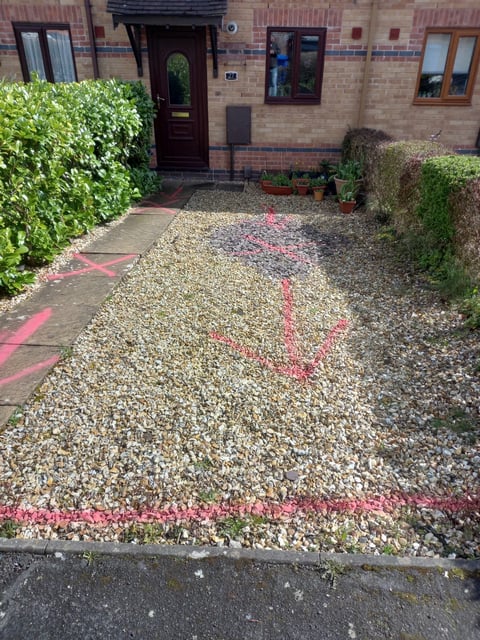 Front courtyard of a brick townhouse with gravel pathway outlined in pink paint, flanked by green hedges and potted plants