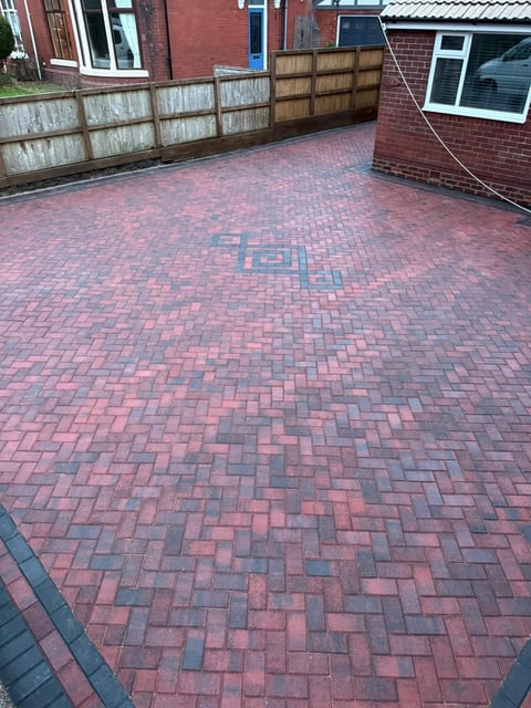 Residential driveway with red and grey herringbone patterned brick pavers, flanked by wooden fence and brick buildings