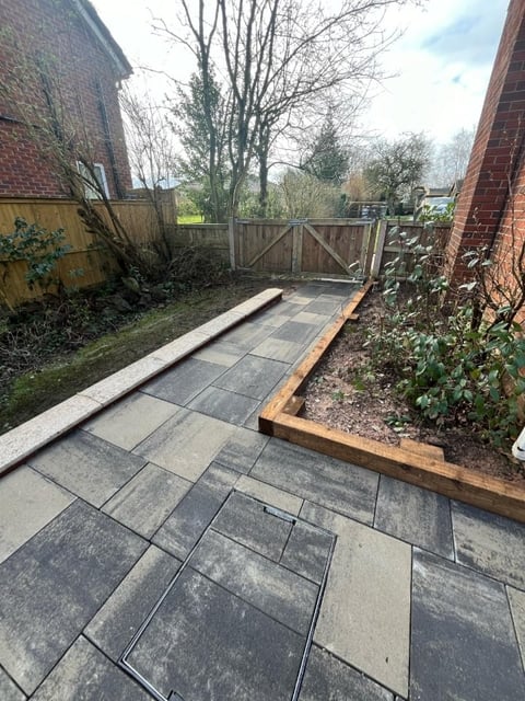 Backyard patio with gray slate tiles, wooden raised garden bed, and gate leading to overgrown yard between brick buildings