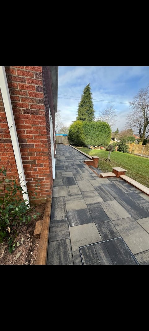 Modern paved patio extending from red brick house with landscaped garden, green hedges, and lawn in background under blue sky
