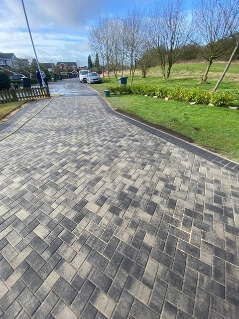 Paved residential driveway with herringbone pattern leading to houses, lawn, and bare trees on a sunny day
