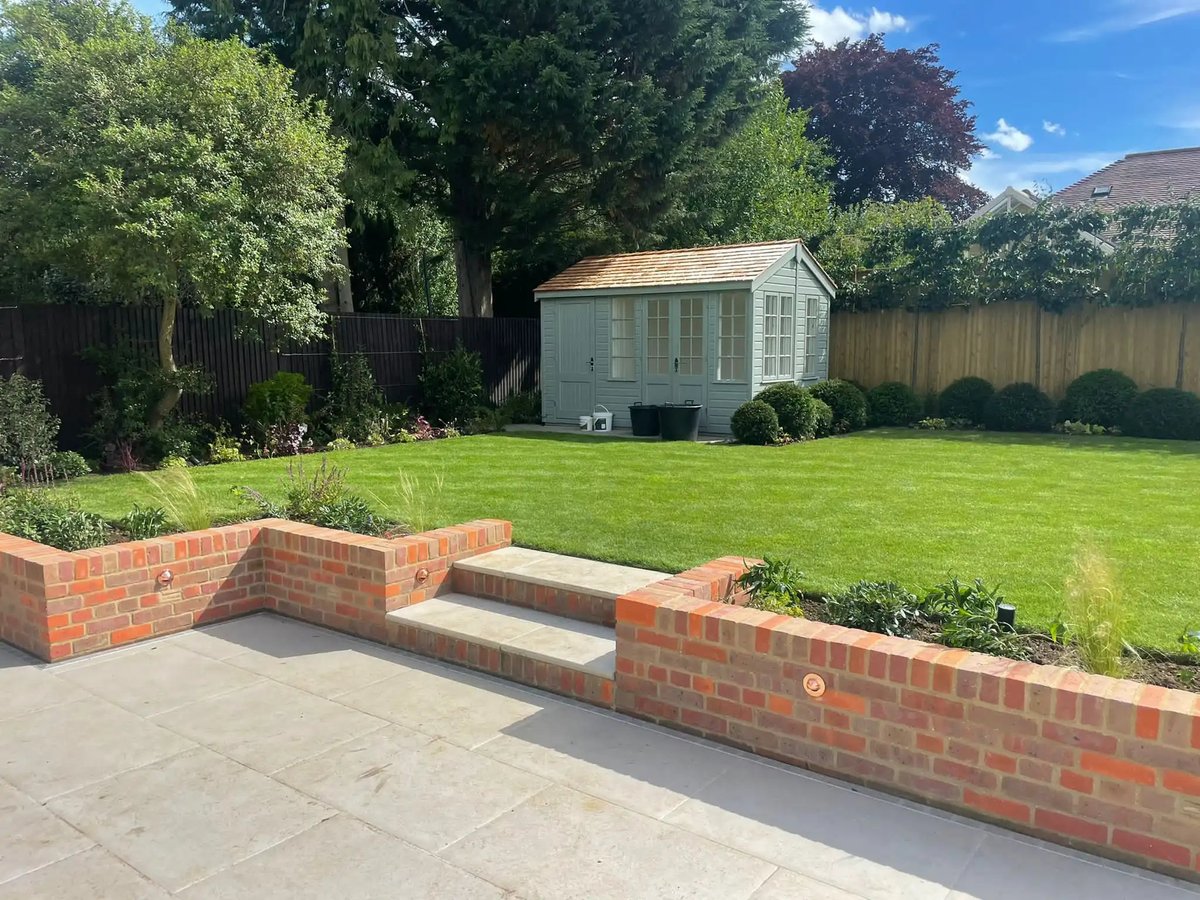 Backyard garden with brick raised beds, patio, lush green lawn, and a small shed surrounded by tall trees and fencing