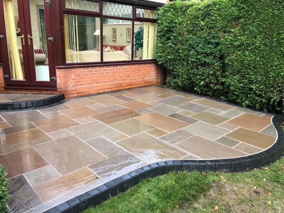 A curved patio with stone paving in shades of brown and tan, bordered by dark edging, adjacent to a brick house with ivy-covered walls and large windows